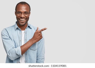 Happy delighted male manager involved in advertising company`s product, indicates with fore finger at blank copy space, wears denim shirt, has positive smile. Advertisement and ethnicity concept - Powered by Shutterstock