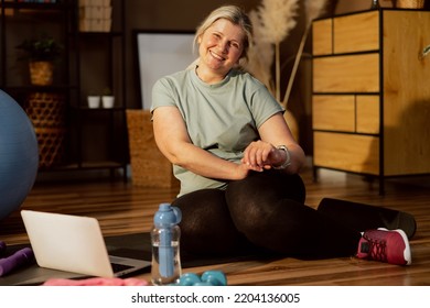 Happy Delighted Older Woman Wearing Sportswear Looking At Camera With Smartwatch On Hand Sitting On Yoga Mat Looking At Camera. Laptop Bottle With Water And Dumbbells On Floor.