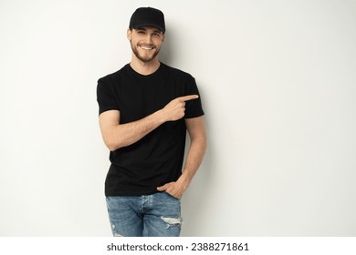 Happy delivery guy employee man wearing black cap t-shirt uniform workwear work as dealer courier pointing aside indicate on area isolated on white background. Service concept