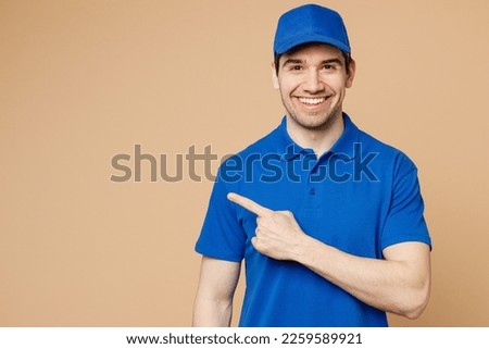 Happy delivery guy employee man wearing blue cap t-shirt uniform workwear work as dealer courier pointing index finger aside indicate on area isolated on plain light beige background. Service concept