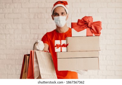 Happy delivery man in medical face mask, Santa hat, protective glove carry shopping bags, cardboard gift boxes with red bow. Young man courier in face mask deliver Christmas presents. Winter sale mall - Powered by Shutterstock