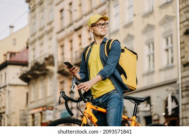 A happy delivery man is sitting on a bicycle and texting on his smartphone. He is carrying a yellow backpack for delivery on his shoulders and cap. He is looking around.
