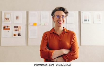 Happy designer smiling at the camera while standing with her arms crossed. Mature businesswoman with eyeglasses working in a creative office. - Powered by Shutterstock