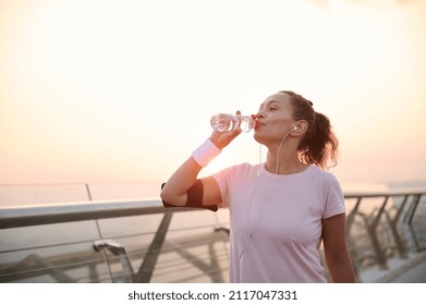 Happy determined athlete, beautiful Hispanic woman wearing pink t-shirt, smartphone holder and terry wristbands stands on a city bridge and drinks water, resting after early morning jog at sunrise - Shutterstock ID 2117047331