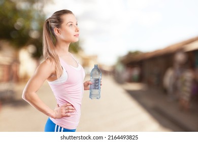 Happy Determined Athlete, Beautiful Woman Stands On A City And Drinks Water, Resting After Early Morning Jog At Sunrise