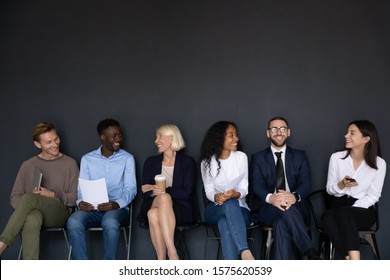 Happy different generations mixed race business people sitting on chairs in row line, isolated on black background with above copyspace. Smiling diverse job seekers chatting, waiting for interview.