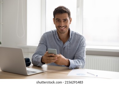 Happy Digital Gadgets User Man Using Smartphone, Laptop At Home Workplace Head Shot Portrait. Young Handsome Business Man Holding Mobile Phone, Sitting At Computer, Looking At Camera