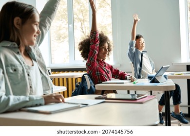 Happy diligent smiling multiethnic diverse schoolchildren having lesson at elementary stem class sitting at desks using gadgets. Schoolkids raising hands answering to questions at workplace.