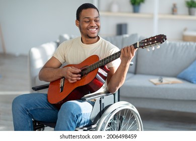 Happy Disabled Black Guy In Wheelchair Playing Guitar, Creating Music In Living Room. Handicapped African American Man Giving Domestic Concert Indoors. Stay Home Activities During Covid Lockdown