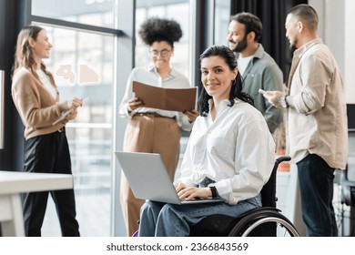 happy disabled businesswoman in wheelchair using laptop near blurred multiethnic colleagues - Powered by Shutterstock