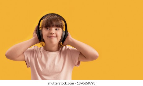 Happy disabled girl with Down syndrome in headphones listening to music and smiling aside while posing isolated over yellow background. Front view. Web Banner