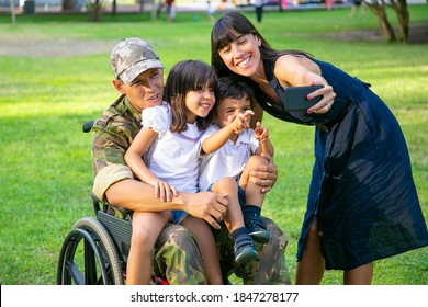 Happy Disabled Retired Military Man Holding Kids In Arms While His Wife Taking Family Selfie On Cell Phone. Veteran Of War Or Returning Home Concept