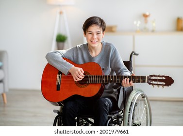 Happy disabled teen boy in wheelchair playing guitar, using musical instrument indoors. Cheerful handicapped adolescent creating music, performing domestic concert. Teenage hobbies and pastimes