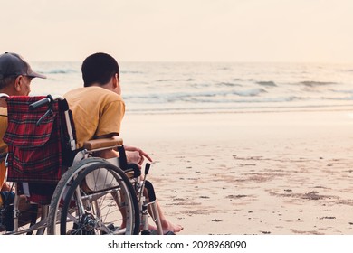 Happy Disabled Teenage Boy On Wheelchair Playing And Relaxing With Parent, Activity Outdoors With Father On The Beach Background, People Having Fun And Diverse Family Concept.