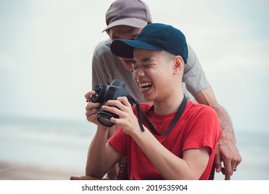 Happy Disabled Teenage Boy Smile Face On Wheelchair Holding A Camera With Father, Activity Outdoors With Family On The Beach Background, People Having Fun And Diverse People Concept.