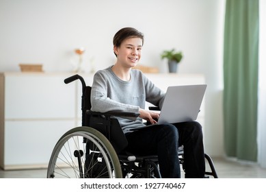 Happy Disabled Teenager In Wheelchair Using Laptop Computer For Online Studies At Home. Positive Handicapped Teen Boy Communicating With Teacher Remotely, Having Webinar Indoors