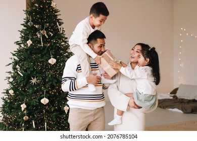 A Happy Diverse Asian Family Celebrates Christmas. Parents With Children Give Gift Boxes Decorate The Christmas Tree And Prepare For The New Year Holiday In A Decorated House. Selective Focus