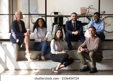 Happy Diverse Business Team People Sit Together On Stairs In Modern Office, Smiling Multiracial Professional Startup Staff Colleagues Group Looking At Camera At Informal Corporate Meeting, Portrait