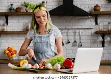 Happy diverse caucasian young teenage girl stylish woman with colorful hair tattoo cooking breakfast, preparing food, tasty dinner salad at home kitchen while using laptop with online tutorial recipe - Powered by Shutterstock