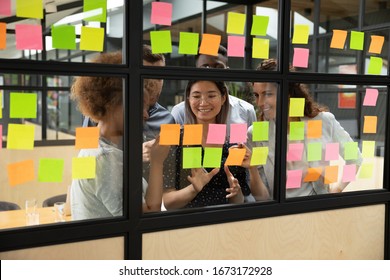 Happy diverse colleagues planning new project, writing creative ideas on stickers on glass board at scrum briefing, successful employees team looking at sticky papers, brainstorming at meeting
