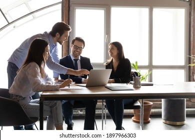 Happy diverse colleagues working on project together, using laptop, sitting at table in modern office, team leader coach training staff, pointing at screen, smiling coworkers discussing strategy - Shutterstock ID 1897966303
