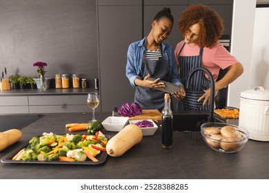 Happy diverse couple in aprons preparing meal, using tablet in kitchen, copy space. Food, cooking, togetherness, relationship, communication, recipe, lifestyle and domestic life, unaltered. - Powered by Shutterstock