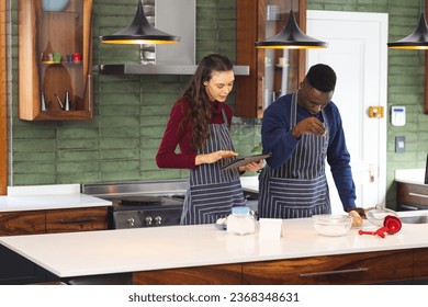 Happy diverse couple baking together in kitchen, using tablet at home, copy space. Lifestyle, togetherness, relationship, baking, recipe, communication and domestic life, unaltered. - Powered by Shutterstock