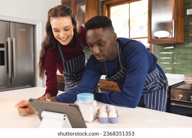 Happy diverse couple baking together in kitchen, using tablet at home. Lifestyle, togetherness, relationship, baking, recipe, communication and domestic life, unaltered. - Powered by Shutterstock