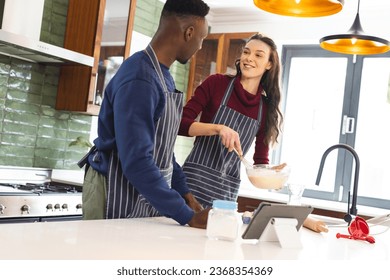 Happy diverse couple baking together in kitchen, using tablet at home. Lifestyle, togetherness, relationship, baking, recipe, communication and domestic life, unaltered. - Powered by Shutterstock