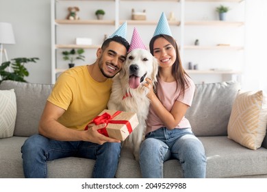 Happy diverse couple celebrating their pet dog's birthday, sitting on sofa with gift box, wearing festive hats at home. Adorable golden retriever having b-day with owners, enjoying party