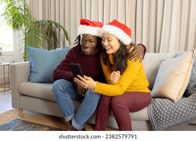 Happy diverse couple in christmas hats having smartphone video call at home, embracing. Communication, christmas, celebration, togetherness, domestic life and lifestyle, unaltered. - Powered by Shutterstock