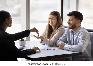 Happy diverse couple of clients getting key from African female realtor, buying house, flat, apartment, sitting at table with paper selling agreement, smiling, laughing, celebrating new ownership - Powered by Shutterstock