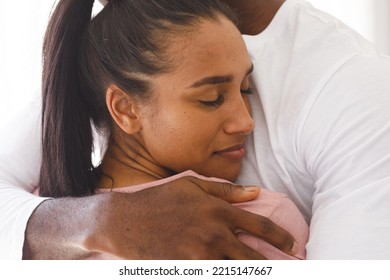 Happy Diverse Couple Embracing By Window At Home, Woman Smiling. Happiness, Domestic Life, Love And Inclusivity Concept.