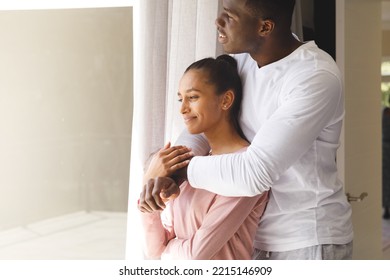 Happy Diverse Couple Embracing And Looking Out Of Window Smiling At Home. Happiness, Domestic Life, Love And Inclusivity Concept.