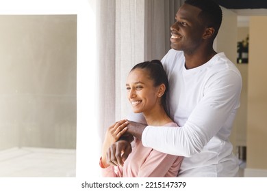 Happy Diverse Couple Embracing, Looking Out Of Window And Smiling At Home. Happiness, Domestic Life, Love And Inclusivity Concept.