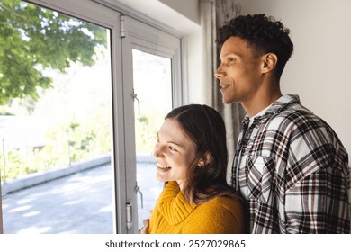 Happy diverse couple embracing and looking through window at home, copy space. Expression, leisure, love, togetherness and domestic life unaltered. - Powered by Shutterstock