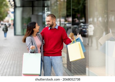 Happy Diverse Couple With Gift Bags And Takeaway Coffee Window Shopping On Street Outside Big City Mall. Cheerful Millennial Spouses Walking Near Supermarket Together, Feeling Excited