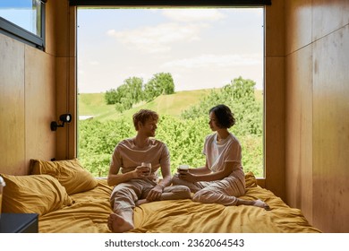 happy diverse couple holding cups of coffee and sitting on bed next to window with beautiful view - Powered by Shutterstock