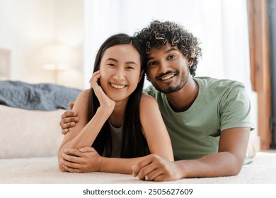 Happy, diverse couple lying on the living room floor, smiling and enjoying time together in a cozy setting with natural light. Celebrating multicultural love and togetherness - Powered by Shutterstock