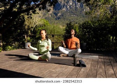 Happy diverse couple practicing yoga meditation sitting on deck in sunny garden, copy space. Yoga, lotus position, meditation, togetherness, relaxation, nature and heathy lifestyle, unaltered. - Powered by Shutterstock