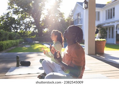 Happy diverse couple practicing yoga meditation sitting on deck in sunny garden, copy space. Yoga, lotus position, meditation, togetherness, relaxation, nature and heathy lifestyle, unaltered. - Powered by Shutterstock