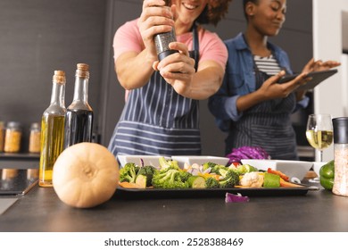 Happy diverse couple preparing meal, using tablet and seasoning vegetables in kitchen. Food, cooking, togetherness, relationship, communication, recipe, lifestyle and domestic life, unaltered. - Powered by Shutterstock