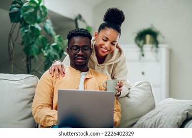 Happy diverse couple relaxing at home and using laptop together. Smiling african american husband and hispanic wife spending time together at home and browsing internet. Hugging and drinking coffee. - Powered by Shutterstock