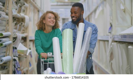 Happy Diverse Couple With Shopping Cart Buying Materials For Redecoration In DIY Store. African Boyfriend And Caucasian Girlfriend Pushing Trolley Purchasing Goods In House Improvement Store