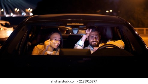 Happy Diverse Couple Sit In Car, Listen To Music And Dancing. View Through Windscreen Of Woman And Man On Date In Car Having Fun And Dancing Late At Night