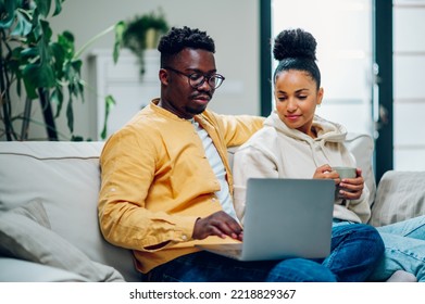 Happy Diverse Couple Sitting And Relaxing On A Couch And Using Laptop Together. Focus On A Smiling African American Husband While Spending Time With His Hispanic Wife At Home And Browsing Internet.