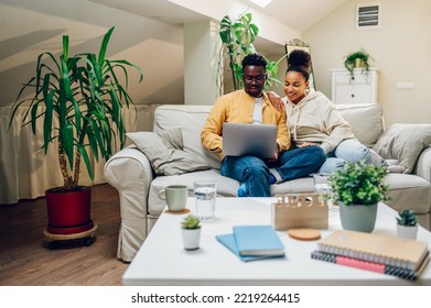 Happy Diverse Couple Sitting And Relaxing On A Couch And Using Laptop Together. Smiling African American Husband And Hispanic Wife Spending Time Together At Home And Browsing Internet. Copy Space.