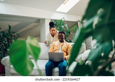 Happy Diverse Couple Sitting And Relaxing At Home And Using Laptop Together. Smiling African American Husband And Hispanic Wife Spending Time Together At Home And Browsing Internet. Copy Space.