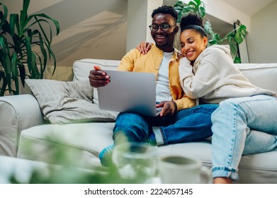 Happy Diverse Couple Sitting And Relaxing On A Couch And Hugging While Using Laptop Together. Smiling African American Husband And Hispanic Wife Spending Time Together At Home And Browsing Internet.