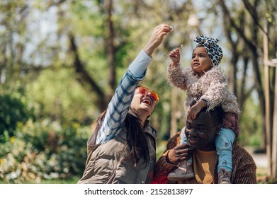 Happy Diverse Family Of Three Spending Their Free Time In The Park And Enjoying Beautiful Sunny Day Outside. Multiracial Parents And Their Cute Little Daughter Having Fun Together.